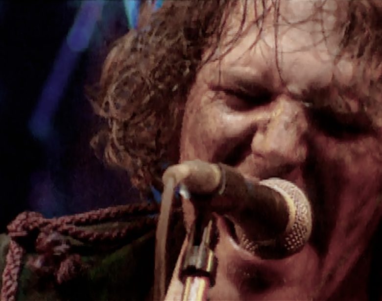 Close up photo of Tim Smith of Cardiacs from live video, singing on microphone